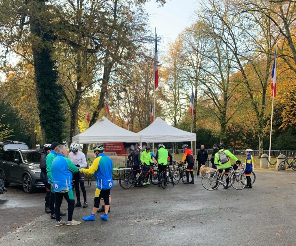 Plusieurs cyclistes vétus de vêtements colorés et fluorescents sont rassemblés devant des barnums à l'entrée de la Clairière de l'Armistice à Compiègne.